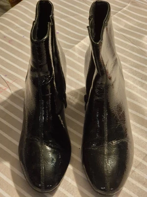 Svarta lackade stövletter med klack - Säljer ett par svarta stövletter i glansigt lackmaterial med markerade sömmar. Skorna har en medelhög blockklack och dragkedja på insidan. Perfekta för dig som vill ha en edgy och trendig look.