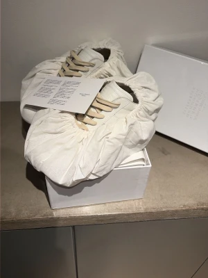 Vita sneakers från Maison Margiela - Stilrena vita sneakers från Maison Margiela i mjukt läder med beige snörning och klassisk gummisula. Snygg minimalistisk design som passar till det mesta. Kommer med originalkartong och skyddande tygpåsar.