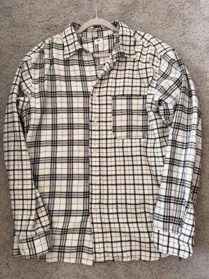 Flannel Vit - Klassisk långärmad skjorta från Uniqlo i vitt med svart rutmönster. Skjortan har en bröstficka, knappar framtill och är tillverkad i mjuk bomull. Perfekt för en avslappnad och stilren look.