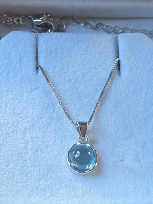 Silverhalsband med blå sten - Snyggt halsband i silver med en rund, blå sten som hänge. Kedjan är tunn och stilren, och stenen har en glansig finish som fångar ljuset. Perfekt för dig som gillar minimalistiska smycken med en färgklick.                          Detaljer: Huvudmaterial: Zinklegering Sten: Rund, slät Cabochon-sten (Ljusblå/ Himmelsblå) Färg: Silvertonad metall Hänges storlek: Ca 1 cm diameter Kedjelängd: 41 - 50 cm Skick: Nytt 