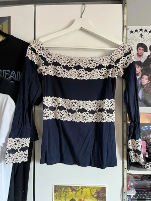 Marinblå intimissimi offshoulder topp med spets - Trendig marinblå offshoulder topp med långa ärmar och breda vita spetsdetaljer vid halsringning, ärmslut och över bröstet. Snygg croppad passform som ger en romantisk vibe. Perfekt för dig som vill sticka ut lite extra.
