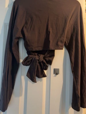 Brun croppad topp med knyt bak - Snygg brun croppad topp från lager 157 med långa ärmar och ett knyt i ryggen. Aldrig använt.