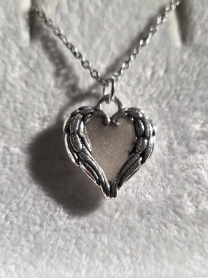 Superfint halsband med hjärthänge och vingar - Unikt halsband med ett hjärtformat hänge inramat av silverfärgade vingar och mitten färgat med vitt och svagt glitter. Hänget har en glödande effekt och lyser blått i mörker. Kedjan är tunn och silverfärgad i rostfritt stål. Perfekt accessoar för dig som gillar detaljer med lite magisk känsla. 🌸✍️🏻// Handgjord & skapad av mig, tillverkat med högkvalitativt självlysande pigment för ett tydligt sken och lång livslängd. Kedjan och hänget tål vatten och missfärgas inte.