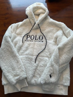 Vit fluffig hoodie från Polo Ralph Lauren - Säljer en supermjuk och fluffig vit hoodie från Polo Ralph Lauren med stor broderad logga på bröstet och klassisk huva med snören. Hoodien har en känguruficka och ribbade muddar. Perfekt för dig som vill ha en cozy och stilren look.