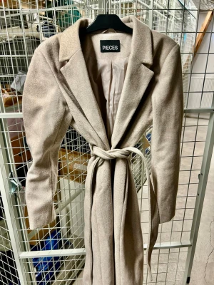 Beige kappa från PIECES med knytskärp - Stilren beige kappa från PIECES med klassisk krage och knytskärp i midjan. Kappan har en rak passform. Perfekt för dig som vill ha en enkel men snygg look till hösten eller våren.