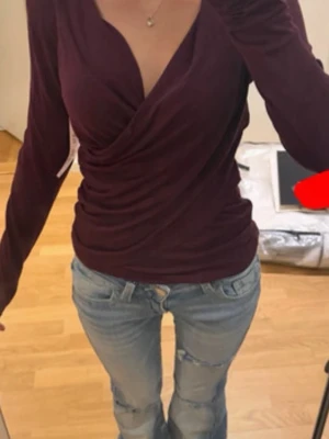 Mörklila omlottopp med v-ringning - Snygg mörklila omlottopp med v-ringning och långa ärmar. Toppen har en figurnära passform och är tillverkad i ett mjukt material som sitter skönt mot kroppen. Perfekt att styla med jeans eller kjol för en trendig look.