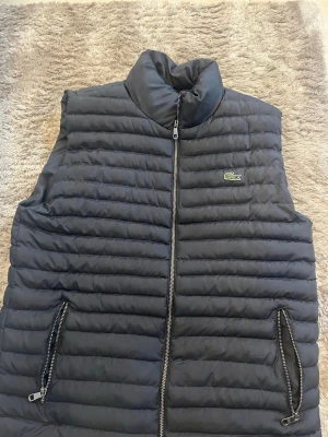Mörkblå dunväst från Lacoste - Snygg mörkblå dunväst från Lacoste med quiltad design och hög krage. Västen har två fickor med dragkedja och klassisk Lacoste-logga på bröstet. Insidan är grön med stort Lacoste-tryck och små krokodiler. Perfekt för lager på lager och streetwear.