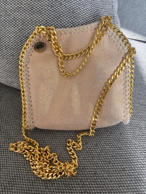 Stella McCartney glitter  rosa/Beige - Supersnygg rosa handväska från Stella McCartney med kedjerem i guld. Den saknar en magnetisk knapp, därav priset. Annars ser den ut att vara i nyskick.Väskan är i veganskt mockaimitationsmaterial och har magnetlås samt innerfack. Jag köpte den här på Plick.