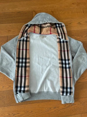 Burrberry zip  - Snygg grå zip-hoodie från Burberry med klassiskt rutigt foder i huvan. Hoodien har dragkedja framtill, två fickor och ribbade muddar vid ärmar och nederkant. Perfekt för en chill och stilren look. Materialet är mjukt och bekvämt, troligen bomullsblandning.super snygg och sitter bea till hösten 