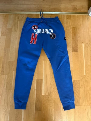 Blå mjukisbyxor från Hoodrich - Snygga blå mjukisbyxor från Hoodrich med vita och röda tryck framtill, elastisk midja med dragsko och muddar vid bensluten. Byxorna har flera coola patches och loggor på framsidan för en streetwear-look. Perfekta för en avslappnad stil.