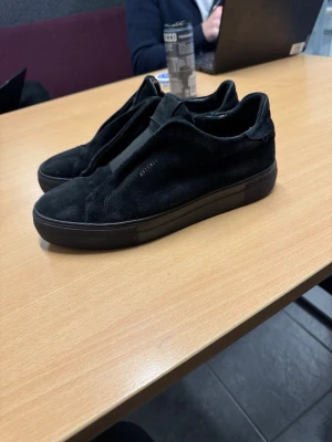 Svarta slip-on sneakers från Axel Arigato - Stilrena svarta sneakers från Axel Arigato i mjuk mocka med diskret logga på sidan i storlek 41 och i bra skick. Slip-on-modell utan snörning och med elastiska paneler för enkel påtagning. Tjock svart sula ger en clean look som passar till det mesta. Lite slitage på mockan på höger yttersida där även lite av texten har blivit lite mindre synlig (se sista bilden). Annars är har dom ett skick på 7/10.