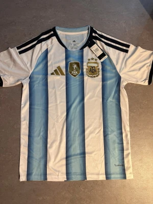 Argentina matchtröja Adidas blåvit - Säljer en officiell Argentina matchtröja från Adidas med klassiska blå och vita vertikala ränder. Tröjan har svarta detaljer på axlar och ärmslut, samt broderade FIFA- och AFA-emblem på bröstet. Tillverkad i lätt och ventilerande material, perfekt för fotboll eller träning.