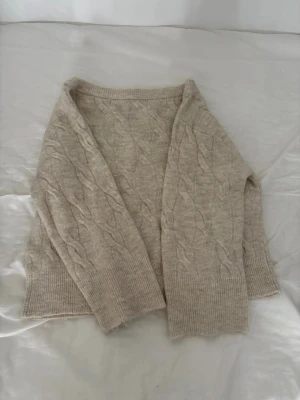Beige kabelstickad tröja off shoulder - Mysig beige kabelstickad tröja med rund halsringning och ribbade muddar vid ärmslut och nederkant. off shoulder. Tröjan har ett klassiskt flätmönster och är perfekt för lager på lager under kyliga dagar. Passar till både jeans och kjol för en avslappnad look.