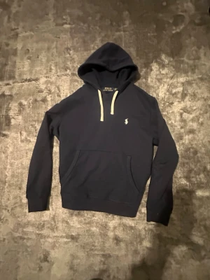 Ralph Lauren Hoodie - Snygg mörkblå hoodie från Polo Ralph Lauren med vit dragsko och klassisk logga broderad på bröstet. Hoodien har känguruficka framtill och ribbade muddar vid ärmar och nederkant. Perfekt för en avslappnad och stilren look.