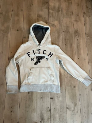 Vit hoodie från Abercrombie & Fitch - Snygg vit hoodie från Abercrombie & Fitch med stor huva och grått innerfoder. Framtill står det 'FITCH NY' i svart text och ett broderat vinge-motiv. Hoodien har känguruficka och ribbade muddar. Perfekt för en avslappnad och trendig look. Något nopprig i tyget.