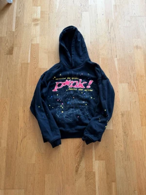 Svart spider hoodie med tryck och stjärnor - Cool svart hoodie med huva och färgglatt tryck framtill där det står 'nevermind the spider pink! heres the slime'. Motiv av spindelnät och små gula stjärnor på både fram- och baksida samt en stor gul stjärna på huvan. Perfekt för dig som gillar unik streetwear.
