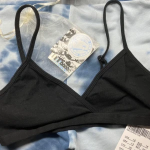 Svart topp/BH från Brandy Melville - En stilren svart BH från Brandy Melville med tunna, justerbara axelband och mjuk resår under bysten. Tillverkad i ett stretchigt och bekvämt material som passar perfekt under alla typer av plagg. Klassisk trekantsform utan vaddering. Med tags kvar. Aldrig använd. Köpt för en vecka sedan. Liten bag med klistermärken följer med.