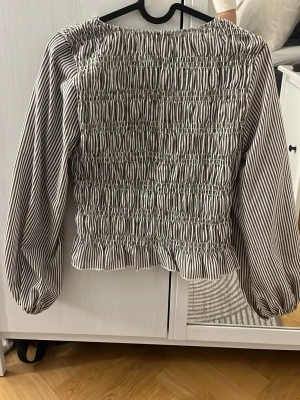 Randig smockad blus med långa ärmar - Säljer en brun vit  randig blus med smockad front och rygg. Blusen har långa puffiga ärmar och rund halsringning. Materialet känns lätt och luftigt, perfekt för en trendig look. Passar till både jeans och kjol.