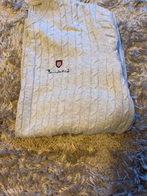 Ljusgrå kabelstickad tröja Bondelid - Snygg ljusgrå kabelstickad tröja från Bondelid med rund halsringning och broderad logga på bröstet. Tröjan har ribbade muddar och är tillverkad i mjukt material, perfekt för lager på lager. Klassisk och stilren design som passar till det mesta.