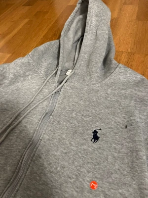 Grå zip hoodie från Polo Ralph Lauren - Snygg grå hoodie med dragkedja från Polo Ralph Lauren. Klassisk design med huva och dragsnören, samt den ikoniska mörkblå polospelaren broderad på bröstet. Tillverkad i mjukt bomullsmaterial och har två fickor framtill. Perfekt för en avslappnad stil.