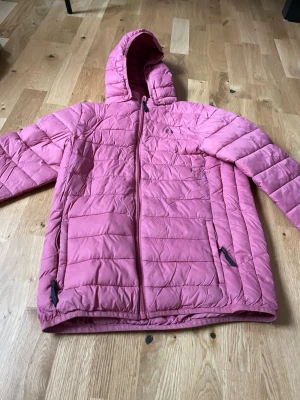 Rosa pufferjacka från Hangten - Rosa pufferjacka från Hangten med huva och dragkedja framtill. Jackan har två fickor med dragkedja och quiltad design. Perfekt för kalla dagar och har en modern, avslappnad look. Materialet är syntet och jackan är lätt vadderad.