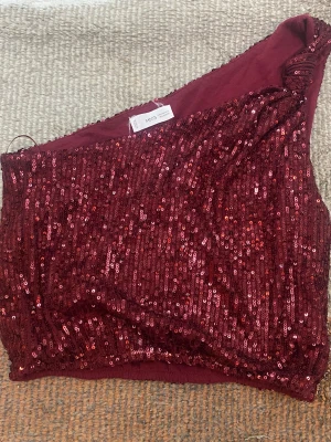 Vinröd paljett oneshoulder topp från Mango - Snygg vinröd oneshoulder topp från Mango, täckt med glittriga paljetter. Perfekt för dig som vill sticka ut och gillar att bära något unikt. Toppens asymmetriska form och paljettmaterial ger en cool och trendig look. Storleken ligger mellan XS och S, använd endast 2 ggr.