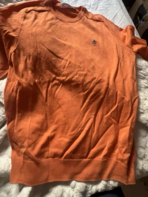 Orange stickad tröja från Morris - Snygg orange stickad tröja från Morris i mjuk merinoull. Tröjan har rund halsringning, ribbade muddar och en liten broderad logga på bröstet. Perfekt för dig som vill ha en färgklick i garderoben.