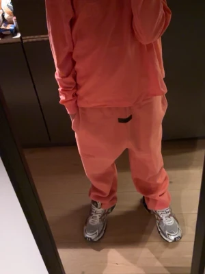 Korallfärgat loungewear-set - Tracksuit ifrån essentials kan även få en hoodie istället för sleeve.  HOODIE STRLK S SLEEVE XS BYXOR S.!!!!! Mysigt loungewear-set i korallfärg med långärmad tröja och matchande byxor. Setet har en avslappnad passform och är perfekt för chill dagar hemma. Materialet ser mjukt och bekvämt ut, och byxorna har resår i midjan. 