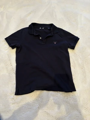 Marinblå pikétröja från GANT - Klassisk marinblå pikétröja från GANT med korta ärmar och krage. Tröjan har två knappar vid halsen och den ikoniska GANT-loggan broderad på bröstet. Perfekt till jeans eller shorts för en clean och stilren look.