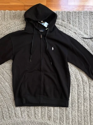 Svart hoodie från Polo Ralph Lauren - Svart hoodie från Polo Ralph Lauren med dragkedja framtill, justerbar huva och fickor på magen. Klassisk vit logga broderad på bröstet. Tillverkad i mjuk bomull för skön känsla och stilren look. Perfekt för dig som gillar enkel och tidlös design. Helt oanvänd 