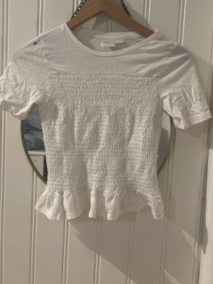 Vit smockad topp med volangkant - Söt vit topp med smockad midja och volangkant nedtill. Kortärmad och rund halsringning, perfekt för en fräsch och enkel look. Tillverkad i mjuk bomull som känns skön mot huden.