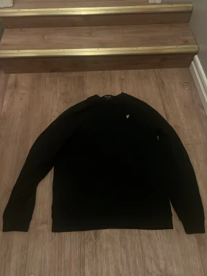 Svart tröja från Lyle & Scott - Klassisk svart tröja från Lyle & Scott med rund halsringning och den ikoniska gula logon broderad på bröstet. Tröjan har lång ärm och en enkel, stilren design som passar till det mesta. Perfekt basplagg för garderoben.