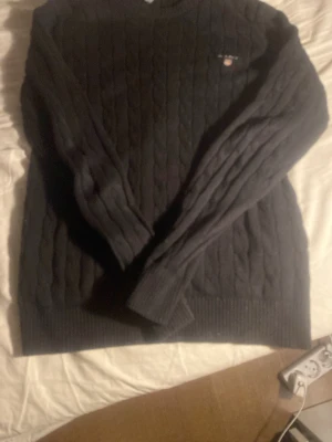 Mörkblå kabelstickad tröja GANT - Kabelstickad mörkblå tröja från GANT med rund halsringning och diskret logga broderad på bröstet. Tröjan har långa ärmar och ribbade muddar vid ärmslut och nederkant. Perfekt för dig som gillar klassisk och stilren look.