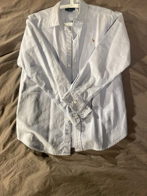 Ljusblå skjorta från Ralph Lauren - Klassisk ljusblå skjorta från Ralph Lauren med button-down krage och broderad logga på bröstet. Skjortan har långa ärmar, knappar framtill och är tillverkad i mjuk bomull. Perfekt för en clean och stilren look.