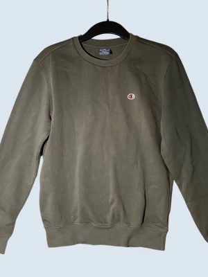 Olivgrön sweatshirt från Champion - Olivgrön crewneck sweatshirt från Champion med liten broderad logga på bröstet. Klassisk modell med rund halsringning, ribbade muddar vid ärmslut och nederkant. Perfekt för en avslappnad och stilren look.