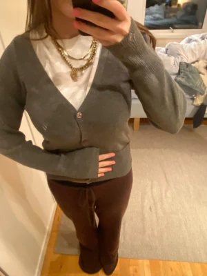 Grå kofta med v-ringning - Mysig grå kofta med v-ringning och knäppning framtill. Långa ärmar och ribbade muddar ger en avslappnad look. Perfekt att slänga över en t-shirt för en enkel och stilren outfit.