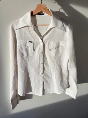 Figursydd skjorta  - Klassisk vit skjorta med långa ärmar, två bröstfickor med lock och knappar framtill. Skjortan har en stilren design med bred krage och diskret logga på ena fickan. 