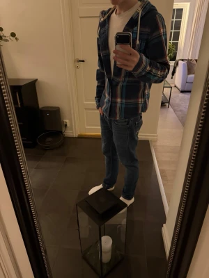 Rutig overshirt från Hollister - Snygg rutig overshirt från Hollister i blått, rött och vitt. Overshirten har huva med dragsko, två bröstfickor med knapp och knäppning framtill. Tillverkad i mjukt bomullstyg, perfekt för lager på lager och en avslappnad streetstil.