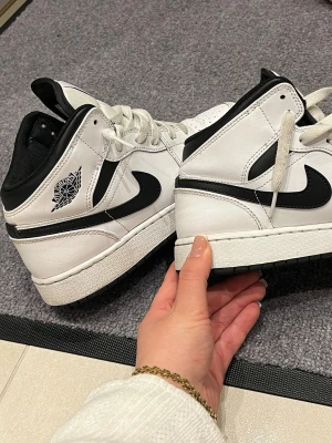 Air Jordan 1 Mid svart/vit - Säljer ett par Air Jordan 1 Mid i svart och vitt läder. Klassisk design med svart swoosh, vita paneler och svart yttersula. Skorna har den ikoniska Air Jordan-loggan på sidan och vita skosnören. Perfekta för dig som gillar streetwear och sneakers med retrovibe.