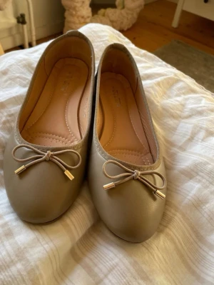 Beige ballerinaskor med rosett - Snygga beige ballerinaskor med rund tå och dekorativ rosett framtill. Skorna har en mjuk innersula och är tillverkade i ett slätt material som ser ut som konstläder. Oanvända och ser helt nya ut.