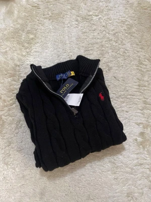 Svart kabelstickad tröja Polo Ralph Lauren - Kabelstickad svart tröja från Polo Ralph Lauren med halv dragkedja och röd broderad logga på bröstet. Tröjan har hög krage och är tillverkad i mjukt material som passar perfekt till kyligare dagar.