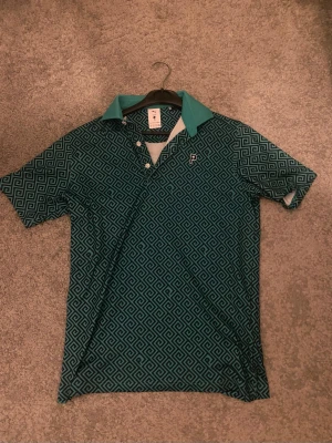 Mönstrad pikétröja från Puma - Snygg blå pikétröja från Puma med grafiskt mönster och kontrasterande turkos krage. Tröjan har korta ärmar, knappar vid halsen och en liten broderad logga på bröstet. Perfekt för en sportig och trendig look.