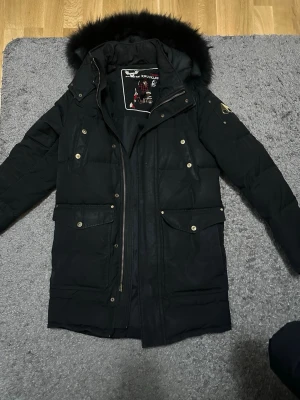 Svart parkas Moose Knuckles med pälskrage - Svart jacka från Moose Knuckles med avtagbar pälskant på huvan, flera fickor med knappar och dragkedja framtill. Klassisk design med guldiga detaljer och varmfodrad insida. Perfekt för kalla dagar.