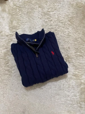 Mörkblå kabelstickad tröja Polo Ralph Lauren - Mörkblå kabelstickad tröja från Polo Ralph Lauren med halv dragkedja och röd broderad logga på bröstet. Tröjan har hög krage och ribbade muddar. Perfekt för dig som gillar klassisk och stilren design.