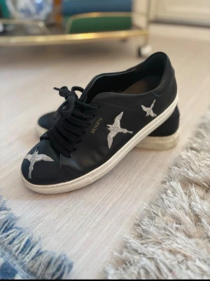 Axel Arigato svarta sneakers med fåglar - Svarta sneakers från Axel Arigato med vita broderade fåglar på sidorna och framtill. Skorna har svart snörning och vit sula i klassisk låg modell. Perfekta för dig som gillar stilrena men ändå unika detaljer.