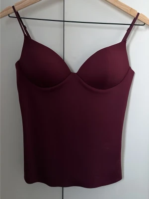 Vinröd body med tunna axelband - Snygg vinröd body från OYSHO med tunna justerbara axelband och vadderade kupor. Tillverkad i ett mjukt och stretchigt material som sitter skönt mot kroppen. Perfekt att styla under en skjorta eller som den är till jeans. Några små fläckar som var där vid köp men går förmodligen att tvätta bort. Endast testad, lappen kvar.
