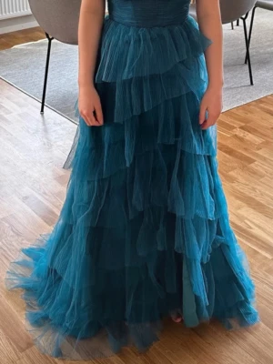 Blå balklänning med volanger - Säljer en blå långklänning med flera lager av volanger i tyll. Klänningen har en figurnära överdel och ett vackert fallande kjolparti som ger en romantisk och elegant look. Perfekt för speciella tillfällen.