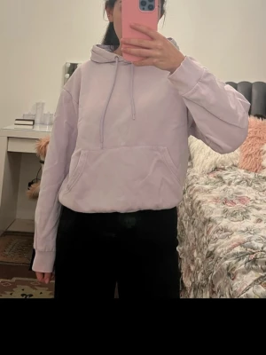 Ljuslila hoodie med huva - En stilren ljuslila hoodie med klassisk huva och dragsnören. Hoodien har en stor magficka framtill och ribbade muddar vid ärmslut och nederkant. Perfekt för en avslappnad look och enkel att matcha med olika outfits.