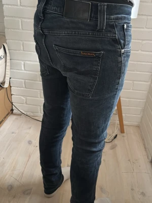 Nudie jeans - Säljer ett par svarta slim fit jeans från Nudie Jeans med klassiska fem fickor och subtila slitningar framtill. Jeansen har en snygg tvättad look, normal midja och är tillverkade i stretchigt denim för extra komfort.