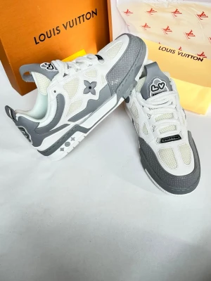 Louis Vuitton vita och grå sneakers - Snygga Louis Vuitton sneakers i vitt och grått med meshpaneler och detaljer i läder. Ikoniska LV-loggor och blommor på sidan samt tungan. Skorna har en chunky sula och coola kontrastdetaljer som ger en modern vibe. Perfekta för dig som vill sticka ut med exklusiva sneakers.